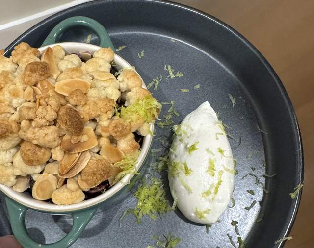 A green mini cocotte of fig crumble under a golden almond
                topping, beside a quenelle of whipped cream scattered with lime
                zest on a dark serving tray.