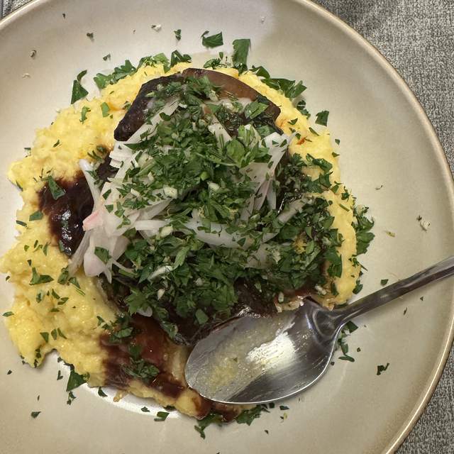 A braised short rib on a bed of yellow parmesan polenta in a wide
                stoneware bowl, buried under chopped parsley gremolata and pink
                pickled shallot, the braising liquid pooling at the edges.
