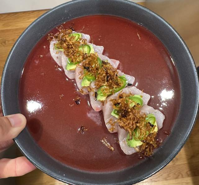 Slices of hamachi laid in a line down a dark bowl of plum-red
                ponzu, topped with avocado, serrano rounds and fried leek.