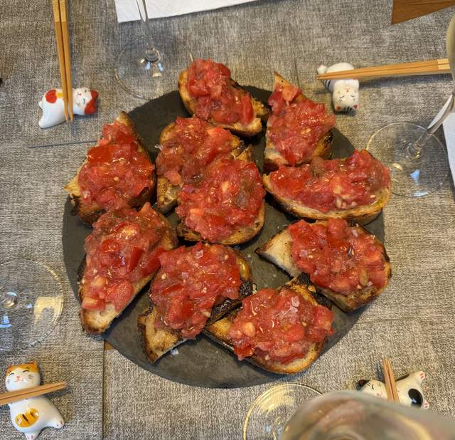 Nine slices of grilled bread heaped with grated tomato and flaky
                salt, in a ring on a round slate board, with wine glasses and
                cat-shaped chopstick rests around it on a grey tablecloth.