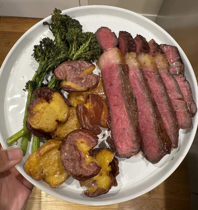 Slices of pink ribeye fanned across a white plate beside crisp
                smashed red potatoes and charred broccolini.