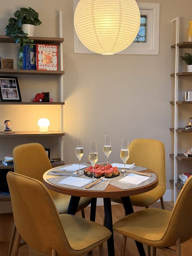 A small round table laid for four with mustard yellow chairs,
                four glasses of sparkling wine, and a slate of pan con tomate
                in the middle, under a paper lantern beside a bookshelf.